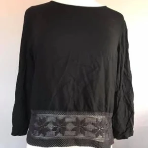 Svart blus med spets från Jacqueline de Yong - Svart blus från Jacqueline de Yong i storlek 38. Blusen har långa ärmar och en rund halsringning. Nedtill finns en bred spetskant med blommönster som ger en snygg detalj. Baksidan har en liten öppning upptill för extra stilpoäng.