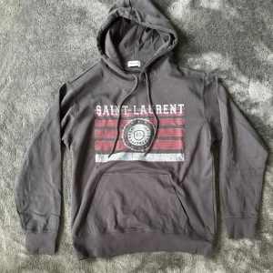Grå hoodie från Saint Laurent - Snygg grå hoodie från Saint Laurent med tryck i vitt och rött på bröstet. Tröjan har huva med snörning, känguruficka och långa ärmar. Materialet är mjuk bomull och passformen är normal. Perfekt för dig som gillar streetwear med exklusiv vibe.