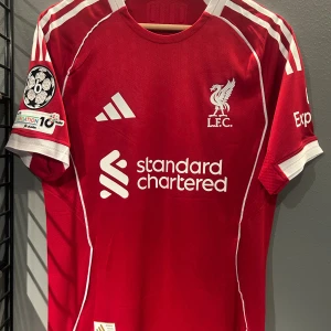 Liverpool Virgil #4 röd Adidas tröja - Liverpool FC matchtröja från Adidas i rött med vita detaljer och tryck. Kortärmad med klassiska vita ränder på axlarna, klubbmärke och sponsortryck på bröstet. På ryggen står det 'Virgil' och nummer 4 i vitt med gul kant. Tillverkad i lätt, ventilerande material. Helt ny liverpool tröja en replika