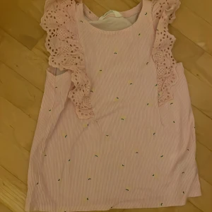 Rosa kortklänning med volang från H&M - Supersöt rosa kortklänning från H&M med tunna vita ränder och små citroner som mönster. Klänningen har breda axelband med broderade volanger som ger en gullig och lekfull vibe. Perfekt för sommarens alla tillfällen!
