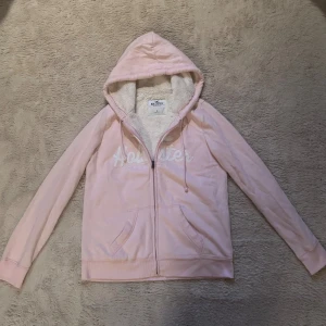 Ljusrosa hoodie från Hollister - Mysig ljusrosa hoodie från Hollister med broderad logga på bröstet. Tröjan har huva med snörning, dragkedja framtill och två fickor. Insidan är fodrad med fluffigt, vitt teddyfoder som gör den extra varm och skön.