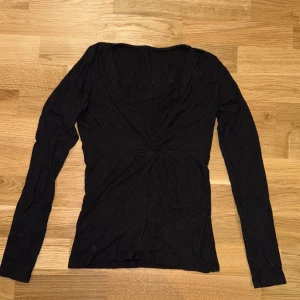 Svart ribbad långärmad topp - Har en liten röd fläck på baksidan Svart långärmad topp med ribbad struktur och snygg twistad detalj framtill. V-ringad hals ger en schysst look och den är figurnära i passformen. Perfekt att styla med jeans eller kjol för en cool vibe.