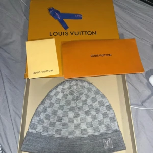 Grå Louis Vuitton beaine - Snygg grå äkta mössa från Louis Vuitton med klassiskt rutigt mönster och LV-logga framtill. Mössan har en uppvikt kant och är tillverkad i mjukt material som håller dig varm. Perfekt accessoar för att lyfta din vinterstil. 