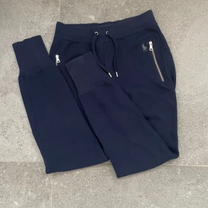 Mörkblå mjukisbyxor från Polo Ralph Lauren - Snygga mörkblå mjukisbyxor från Polo Ralph Lauren med silverfärgade dragkedjor vid fickorna och klassisk logga broderad på framsidan. Byxorna har snörning i midjan och ribbade muddar vid bensluten för extra komfort. Perfekt för chill dagar. Pris kan diskuteras😊!