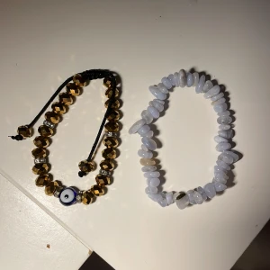 Två armband med pärlor och stenar - Säljer två snygga armband! Ett med guldiga facetterade pärlor, strassdetaljer och ett blått öga, samt ett med ljusblå oregelbundna stenar på elastisk tråd. Perfekta accessoarer för att lyfta din stil.