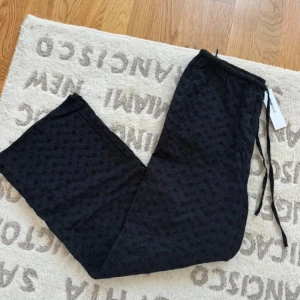 Svarta broderade byxor med snörning - Svarta byxor från Kappahl i storlek S med engelsk brodyr och blommigt mönster. De har elastisk midja med snörning och är tillverkade i bomull. Passformen är loose och benen är raka för en chill vibe.