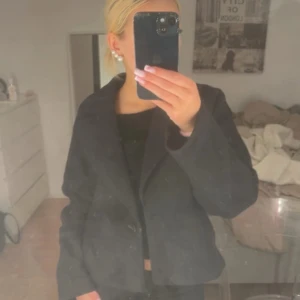 Svart kappa från VILA - Snygg svart kappa från VILA i klassisk modell med krage och två knappar framtill. Jackan har lång ärm och är lätt croppad i passformen, vilket ger en modern vibe. Perfekt att styla med jeans eller kjol för en cool look.