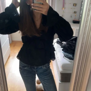 Svart blus med volang - Säljer en svart blus med volangdetaljer och långärmade puffiga ärmar. Blusen har en v-ringad hals och är tillverkad i ett lätt, strukturerat tyg som ger en snygg och trendig look. Perfekt att styla med jeans eller svarta byxor.