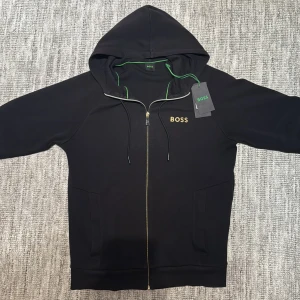 Boss - Svart hoodie från BOSS med gulddetaljer på dragkedjan och broderad BOSS-logga på bröstet. Tröjan har huva med gröna inslag, snörning och två fickor framtill. Tillverkad i mjuk bomull och har en normal passform. Perfekt för dig som gillar stilrena och exklusiva plagg. Helt ny och pris kan diskuteras.