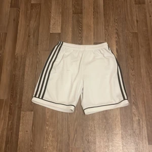 Vita Adidas shorts med svarta ränder - Säljer ett par vita Adidas shorts med klassiska svarta ränder längs sidorna och svart kant längst ner. De har elastisk midja med snörning och är gjorda i mjukt polyester-material. Perfekta för sport eller chill. Adidas-logga på vänster ben.