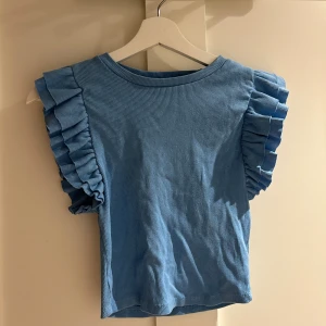 Blå volangblus från Zara - Supersöt blå blus från Zara i ribbad bomull med volangdetaljer på axlarna. Modellen är ärmlös med rund hals. Perfekt för dig som gillar att sticka ut med coola detaljer och mjukt material.