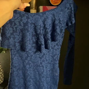 Blommig blå spetsig oneshoulder topp - Säljer en snygg blå topp med blommigt spetsmönster och oneshoulder-design. Toppen har lång ärm på ena sidan och en volangdetalj över bröstet. Perfekt för dig som gillar att sticka ut med coola detaljer och unik form.