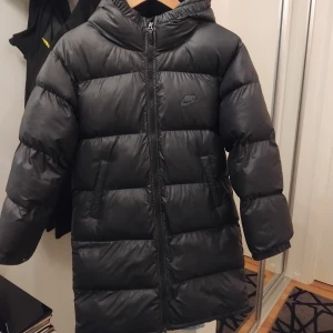 Lång nike jacka  - Säljer en svart pufferjacka från Nike med huva och dragkedja framtill. Jackan har ett stilrent Nike-logga på bröstet och praktiska innerfickor. Den är vadderad och har en längre modell som håller dig varm under kalla dagar. Perfekt till vintern. Jackan är storlek xs men passar även s jag vill sälja den eftersom jag vill köpa en ny jacka använt ett fåtal gånger