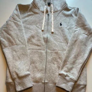 Grå hoodie från Polo Ralph Lauren - Snygg grå hoodie från Polo Ralph Lauren med klassisk broderad logga i marinblått på bröstet. Tröjan har huva med vita snören, hel dragkedja och ribbade muddar. Perfekt för chill dagar och enkel att matcha med jeans eller joggers.