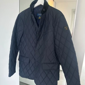 Mörkblå quiltad jacka Polo Ralph Lauren - Snygg mörkblå quiltad jacka från Polo Ralph Lauren med klassisk krage i manchester, broderad logga på ärmen och praktiska fickor framtill. Jackan har både dragkedja och knappar, perfekt för dig som gillar stilren och tidlös design.