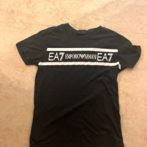 Svart EA7 Emporio Armani t-shirt - Svart t-shirt från EA7 Emporio Armani med vit logga och tryck både fram och bak. Klassisk rund hals och korta ärmar. Snygg och stilren design med raka linjer över bröstet. Perfekt för dig som gillar exklusiva streetwear-plagg.