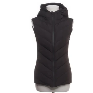 Svart dunväst med huva - Snygg svart dunväst med huva och dragkedja framtill. Västen har ett quiltat mönster i diagonala linjer som ger en cool look. Perfekt att slänga över en hoodie eller tröja när det är kyligt ute. Enkel och stilren design som funkar till många outfits.