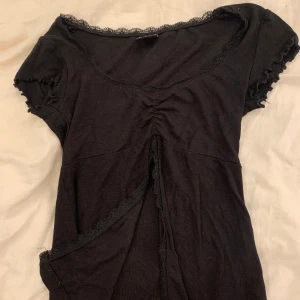 Svart blus med spets från Gina Tricot - Supersöt svart blus från Gina Tricot i storlek XS. Toppen har korta ärmar med volangkant, spetsdetaljer runt halsringningen och framtill, samt en snygg v-ringad form med rynk och omlott-effekt. Perfekt för dig som gillar romantiska detaljer!