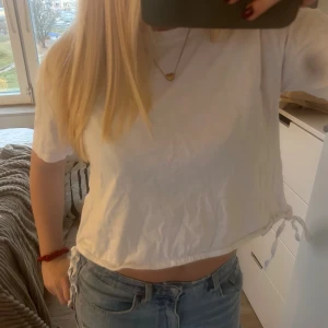 Vit croppad topp med snörning från Zara - Säljer en vit croppad topp från Zara med korta ärmar och snygg snörning i sidorna. Toppen är i mjuk bomull och har en enkel, clean look som är lätt att matcha med jeans eller kjol.