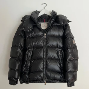 Svart Moncler pufferjacka med huva - Svart pufferjacka från Moncler i fint skick, med stor avtagbar huva och två sidofickor med dragkedja. Jackan har Moncler-logga på ärmen och kraftig dragkedja framtill. Stor i storleken så skulle säga att det är som en storlek S. Bröstvidd: 54 cm, längd: 64 cm, ärmlängd: 61 cm. Perfekt för kalla dagar!
