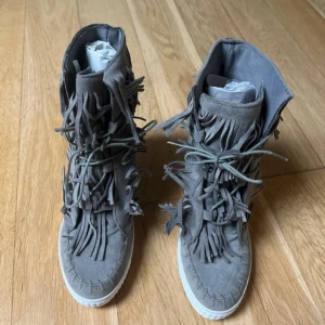 Grå mockaboots med fransar från Vices - Grå boots från Vices i mjuk mocka med coola fransdetaljer runt hela skaftet och framtill. Skorna har snörning.