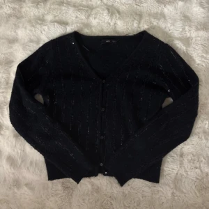 Svart glittrig kofta från Vero Moda - Snygg svart kofta från Vero Moda med glittriga inslag och v-ringning. Koftan har knappar framtill och är långärmad, perfekt att slänga över en topp för en extra touch. Den är stickad och har en klassisk passform.