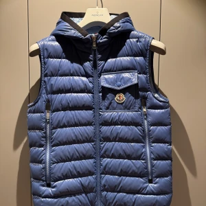 Blå dunväst med huva från Moncler - Snygg blå dunväst från Moncler med quiltad design och huva. Västen har dragkedja framtill, två sidofickor med dragkedja och en bröstficka med knapp och Moncler-logga. Kvitto finns, hör av dig för mer info