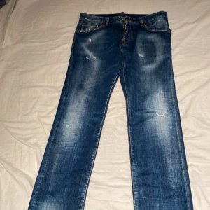 Blå jeans från Dsquared2 - Säljer ett par snygga blå jeans från Dsquared2 med slitna detaljer och klassisk femficksdesign. Jeansen har tydliga kontrastsömmar, knappgylf och Dsquared2-logga på både knapp och etikett. Passformen är rak och materialet är robust denim.