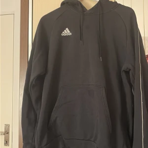 Svart Adidas hoodie XL med huva - Svart hoodie från Adidas i storlek XL med vit broderad logga på bröstet. Tröjan har huva med snörning, stor magficka och vita detaljer längs ärmen. Mjuk insida i fleece och klassisk loose passform, perfekt för chill eller träning.