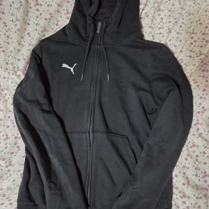 Svart hoodie från Puma med dragkedja - Svart hoodie från Puma med vit logga på bröstet, hel dragkedja och snörning i huvan. Tröjan har långa ärmar och en stor ficka framtill. Mjuk insida i bomullsmix som är skön att ha på sig. Perfekt för chill eller träning.