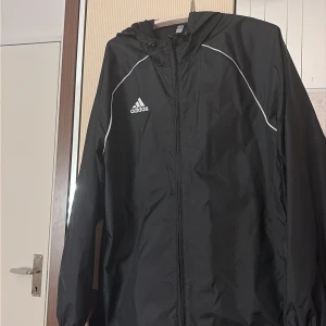 Svart vindjacka från Adidas L - Svart vindjacka från Adidas med vit logga på bröstet och vita detaljer längs axlarna. Jackan har huva med snörning, dragkedja framtill och fickor med dragkedja. Tillverkad i lätt polyester, perfekt för aktiva dagar och sport.