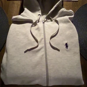 Grå hoodie från Polo Ralph Lauren XS - Snygg grå hoodie från Polo Ralph Lauren i storlek XS. Tröjan har dragkedja, huva med snörning och två fickor fram. Klassisk svart Polo-logga på bröstet. Mjuk bomullskvalitet och långärmad modell, perfekt för chill dagar. Pris går att förhandlas! 