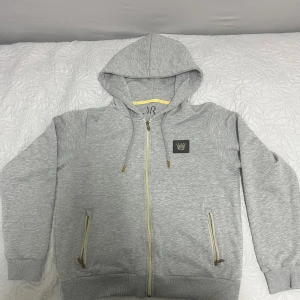 Grå hoodie med gulddetaljer från M.R - Snygg grå hoodie från M.R med gulddetaljer på dragkedjor och snören. Tröjan har huva, två fickor med dragkedja och en svart patch med krona på bröstet. Perfekt för chill dagar och streetstyle.. väldigt bekväm tröja och ganska sällsynt att se denna koftan utomhus.