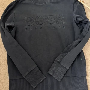 Svart sweatshirt från Hugo Boss - Snygg svart sweatshirt från Hugo Boss med broderad logga framtill. Tröjan har långa ärmar, ribbade muddar och rund halsringning. Perfekt för en stilren och avslappnad look.