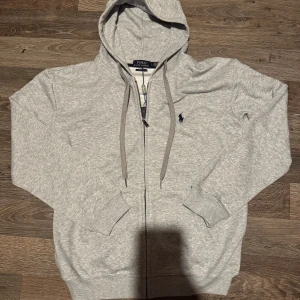 Grå Ralph Lauren zip-hoodie - Snygg ljusgrå hoodie från Ralph Lauren med dragkedja, huva och snörning. Tröjan har den klassiska loggan på bröstet och är tillverkad i mjuk bomull. Perfekt för chill dagar eller när du vill ha en clean och stilren look.