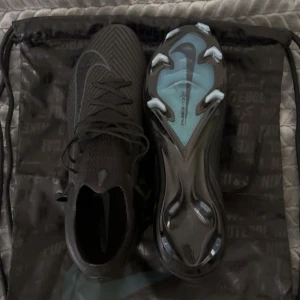 Nike svarta fotbollsskor med snörning Inkluderat PÅSE!! - PRIS KAN DISKUTERAS!! Säljer ett par svarta Nike fotbollsskor med snyggt mönstrad ovandel och ljusblå detaljer på sulan. Skorna har snörning och är tillverkade i syntetmaterial med en tight passform. Sulan har dobbar för gräsplan/konstgräs och en stilren Nike-logga på sidan och nästan oanvända.
