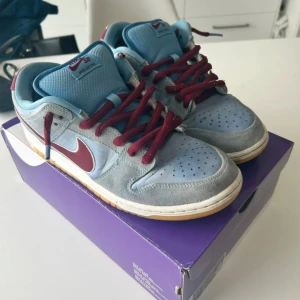 Nike SB Dunk Low PRM blå/vinröd - Nike SB Dunk Low PRM i färgerna valor blue och team maroon. Skorna har vinröda snören, ljusblå mesh på tungan och gråblå mocka på ovandelen. Skorna har slitage under skon men inte så att det läcker in.Swoosh-loggan är vinröd med vit kant, och sulan är vit med gummidetaljer. Perfekt för dig som gillar unika sneakers.
