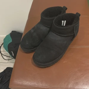 Svarta UGG boots i mocka - Mysiga svarta UGG boots med låg skaft och rund tå. Utsidan är i mjuk mocka och insidan är fodrad med fluffigt material som håller fötterna varma. Klassisk UGG-logga baktill och platt sula för skön komfort. Perfekt för kalla dagar och chill outfits.