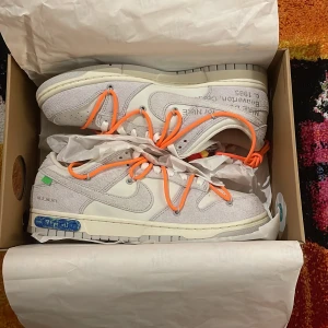 Nike Dunk Low Off-White - Nike Dunk Low Off-White sneakers i ljusgrå mocka och vitt skinn med unika orange snören och detaljer. Snygg text på sidan och coola Off-White detaljer. Kommer i specialdesignad låda med hål och grafiska tryck. Perfekt för dig som vill sticka ut.