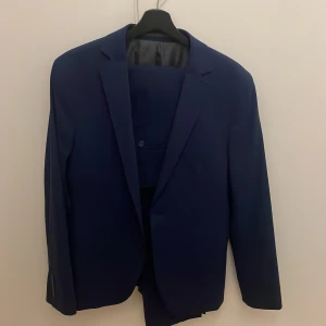 Mörkblå kostym från Dressmann, slim fit - Säljer en stilren mörkblå kostym från Dressmann i storlek 48, slim fit. Kavajen har klassisk passform med enkla knappar och matchande byxor. Perfekt för dig som vill ha en snygg och modern look. Materialet är mjukt och följsamt för extra komfort.