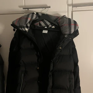 Svart dunjacka från Burberry M - Säljer en svart dunjacka från Burberry med klassiskt rutigt foder i huvan. Jackan har dragkedja, tryckknappar och en tydlig Burberry-patch på ärmen. Perfekt för kalla dagar med sin fluffiga och varma känsla. Stilsäker och tidlös design. Helt ny med avtagbara armar!