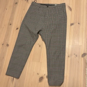 Rutig kostymbyxa från HOPE, storlek 40 40 - Snygga rutig kostymbyxa från HOPE i grått och svart med diskreta röda detaljer. Modellen har tapered ben, fickor fram och bak samt hällor för bälte. Materialet är en mix av polyester och ull med lite stretch för extra komfort.