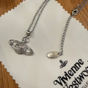Vivienne Westwood halsband i silver - Snyggt halsband från Vivienne Westwood med ikonisk orb-berlock i silverfärgad metall och glittriga stenar som dessutom aldrig är använt! Kedjan är justerbar i olika storlekar och har en liten tag med loggan och kommer med originalförpackning, dustbag och certifikat. Perfekt statement piece för dig som gillar trendiga accessoarer.