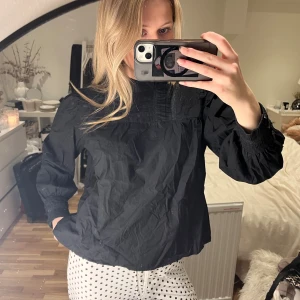 Svart blus med puffärm - Svart blus med puffiga långärmar. Blusen har smock vid ärmslut och diskreta broderade detaljer upptill. Materialet är lätt. Passformen är normal och modellen är klassisk med en modern twist.