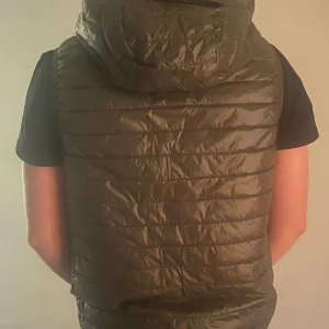 Olivgrön dunväst från Replay - Snygg olivgrön dunväst från Replay med huva och quiltad design. Västen har dragkedja framtill med Replay-logga och ett Replay-märke på bröstet. Perfekt att slänga över en hoodie för extra värme under hösten eller vintern. Storlek M.