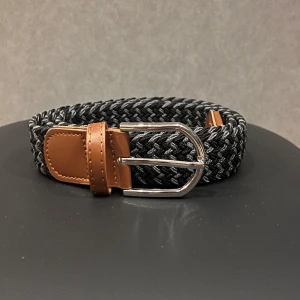 Svart och grått flätat bälte - Snyggt flätat bälte i svart och grått med bruna skinn-detaljer vid spännet och ändarna. Spännet är i blankt silverfärgat metall och bältet har en modern, stilren look som passar till jeans eller chinos. Perfekt accessoar för att lyfta din outfit.
