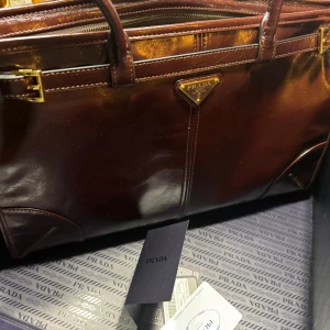 Brun skinnportfölj från Prada - Snygg portfölj från Prada i mörkbrunt, glansigt skinn med guldfärgade metalldetaljer och klassisk trekantig logotyp framtill. Väskan har dubbla handtag, dragkedja upptill och ett rymligt fack med exklusivt mönstrat innerfoder.