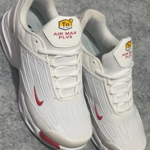 Nike Air Max PLV9 vita sneakers - Nike Air Max PLV9 sneakers i fräsch vit färg med röda detaljer och Tn Air-logga på plösen. Snyggt mönstrad ovandel i syntet och mesh, med klassisk snörning och platt sula. Perfekt för dig som gillar sportig och clean stil.