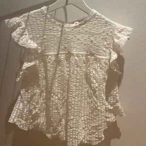 Vit blus med volangärmar och spets - Supersöt vit blus med korta volangärmar och spetsdetaljer. Blusen har ett strukturerat rutmönster och rund halsringning. Luftig och flowy passform, perfekt för ljusa dagar. Materialet känns som bomull och spets, vilket ger en romantisk vibe.