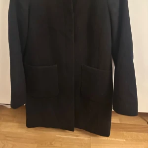 Svart kappa från Oasis XS - Stilren svart kappa från Oasis i storlek XS. Klassisk rak modell med stora fickor framtill och diskret knappstängning. Perfekt för dig som gillar minimalistisk look och vill ha en jacka som funkar till många olika outfits.